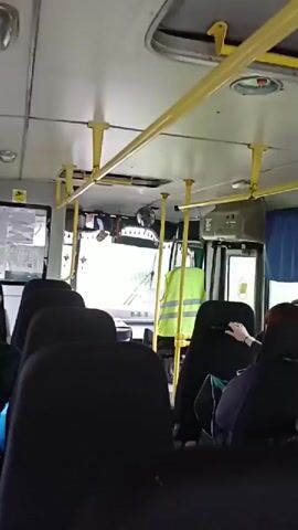 The video shows the interior of a bus with passengers seated. The camera pans from the front of the bus towards the back, revealing rows of black seats. Some passengers are visible, including a woman with braided blonde hair and a person wearing a black cap. Blue curtains are drawn across the windows on one side of the bus. In the front of the bus, a high-visibility yellow vest is hanging. The lighting appears to be natural, suggesting daylight.