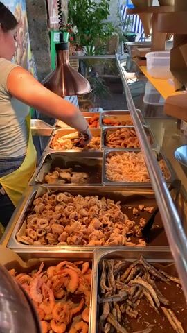 A young woman with her hair pulled back, wearing a light-colored t-shirt and a bright yellow apron, is serving food from a display case. She is using tongs to place items into a small container. The display case is filled with various fried seafood dishes, including calamari rings, shrimp, and small fish. The food is arranged in large metal trays. Behind the woman, there are more food items and some greenery, suggesting an outdoor food stall or market. The scene appears to be brightly lit, possibly indicating daytime. The overall atmosphere seems to be that of a busy food service environment.