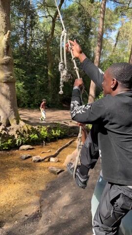A man in black clothing, including pants with "CLINTS" printed on the lower leg, swings on a rope swing over a shallow stream in a wooded area. He is seen holding onto the thick rope, propelling himself forward and backward, with his legs extended during the swing. Another person, partially visible on the right, is helping to push the swing. In the background, a third man stands on a raised bank near a large tree, observing the activity. The scene is outdoors, set in a forest or park with numerous trees, exposed roots, and rocks scattered along the stream bed and banks. The lighting suggests it is daytime, likely sunny. The ground is earthy with patches of green foliage. The location is Poole, United Kingdom.