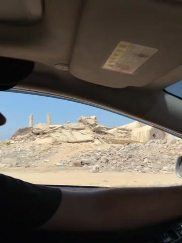 The video shows a view from inside a car. The car is driving through a desolate, rocky, and debris-strewn landscape. In the distance, there are large piles of rubble and what appear to be the remains of buildings, with some concrete structures and pillars visible. The sky is clear and blue. The dashboard and the windshield of the car are visible, with the windshield showing significant cracks. A person's arm and hand are on the steering wheel. The sun visor is also visible, with a yellow sticker on it. The video appears to capture a journey through a damaged or destroyed area.