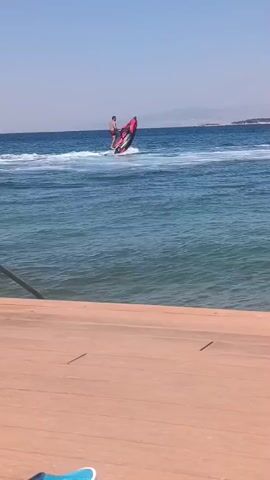 A person is riding a red jet ski on a body of water. The person is standing on the jet ski and appears to be turning. The jet ski is creating a wake behind it. The water is blue and clear. There is a wooden surface in the foreground, possibly a dock or a deck. In the background, there is a landmass with trees and hills, and the sky is clear and blue. The overall scene depicts a recreational activity on a sunny day.