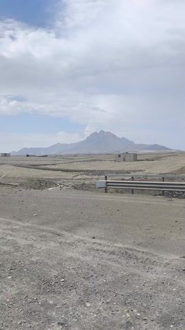 The video depicts a vast, arid landscape, characterized by gray, gravelly terrain under a partly cloudy sky. In the distance, a prominent mountain range stands out against the horizon. Several small, box-like structures are scattered across the plain between the viewer and the mountains. A road runs parallel to the viewer's position, separated by a metal guardrail. An object that appears to be a signpost with an orange tip is angled downward, seemingly damaged or broken. There is no visible human activity or significant movement observed in the video. The scene suggests a remote and desolate environment.