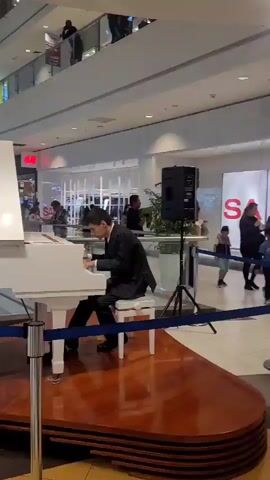 A man in a dark suit and tie is playing a white grand piano on a raised platform in a mall. He is seated on a white stool. To the right of the piano is a black speaker on a stand. Blue ropes cordon off the area around the piano. In the background, several people are visible on upper levels of the mall and walking through the shopping area. The mall appears to be well-lit with shops and escalators visible. In the second clip, a person wearing a white t-shirt and dark pants walks away from the camera, carrying a white shopping bag with a yellow handle. The floor is tiled in a checkerboard pattern of light and dark squares.