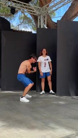 The video shows two young men dancing on a stage outdoors. The man on the left is shirtless and wearing blue shorts and white sneakers. He is facing forward with his left arm raised. The man on the right is wearing a white t-shirt with "STAFF" printed on it in large letters, blue shorts, and white sneakers. He is facing forward and appears to be dancing along with the other man. Both men are smiling and seem to be enjoying themselves. The stage is made of concrete, and there are black backdrops behind the men. A large white screen is visible to the left of the stage. Overhead, there is a metal structure with lights and what appears to be a tree branch. The overall impression is of a casual, outdoor performance or event.