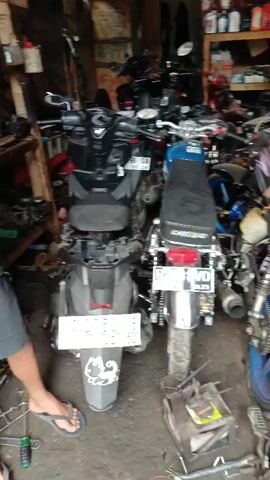 The video shows a cluttered garage or workshop filled with motorcycles. In the foreground, there are three motorcycles parked close together. The motorcycle on the far left is a dark-colored scooter with its rear seat removed, revealing some internal components. Next to it is a blue vintage-style motorcycle with chrome accents and a license plate. To the right of that is another motorcycle with a blue rim and a black seat, partially obscured by the first motorcycle. In the background, more motorcycles are visible, along with shelves stocked with various items, possibly spare parts or fluids. Tools and parts are scattered on the floor around the motorcycles. The environment appears to be indoors, with brick walls and a wooden ceiling. The lighting is dim, suggesting it might be an enclosed space with artificial lighting.