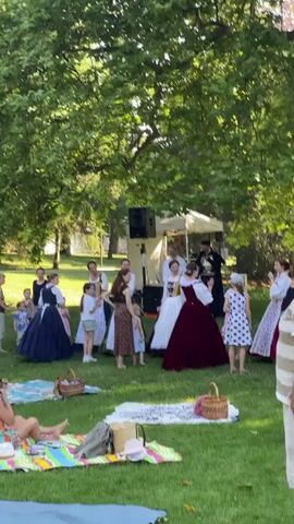This video captures an outdoor event in Stupava, Slovakia, likely a cultural performance or historical reenactment. The scene is set in a park or garden with lush green grass and a large, leafy tree providing shade. A group of people, predominantly women and girls dressed in what appear to be traditional or historical costumes, are gathered around a small stage area.

The costumes vary, with some featuring full, long skirts in dark blues and reds, and others in lighter colors and patterns. One girl wears a white dress with a white bow in her hair, while another is in a polka-dot dress. Some of the adult women are also in period attire, while a few are in more casual, modern clothing, suggesting they might be organizers or audience members.

In the background, a white tent is visible, possibly serving as a backdrop or shelter for performers. A portable speaker system is set up, indicating that music or announcements are part of the event. Several individuals are standing and interacting, some appearing to be performing or rehearsing, while others watch from the sidelines.

In the foreground, several people are seated on picnic blankets, enjoying the event. The sunlight filtering through the trees suggests it is daytime, likely late afternoon given the quality of light. The overall atmosphere appears relaxed and festive, with families present, including young children. The presence of picnic baskets further emphasizes a leisurely outdoor gathering. No visible text is present in the video.