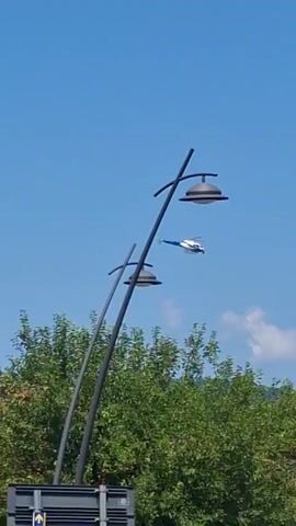 A white and blue helicopter flies across a clear blue sky. In the foreground, two streetlights with curved poles are visible. Lush green trees and a few buildings with red-tiled roofs are also in the scene. A road sign indicating directions to various locations, including "VANGILE 5,1 KM," "CROCI 3,1 KM," and "MASSACIO 8,1 KM," is present, along with a yield sign and a roundabout sign.