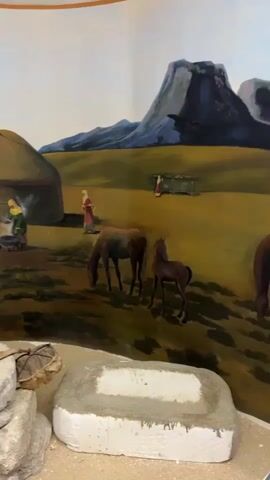 The video shows a mural depicting a nomadic scene with yurts, horses, and mountains. In the foreground, there are various artifacts, including millstones, ropes, and what appears to be a wooden rake or tool. The mural features people tending to a fire outside a yurt, a person in a red dress standing near another yurt, and a camel being attended to. The overall environment depicted in the mural is a grassy plain with a large mountain in the background. The lighting suggests it is daytime.