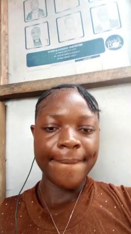 A young Black woman with braided hair and a brown shirt is centered in the frame. She is wearing a thin silver necklace with a small pendant. She is looking directly at the camera with a slight smile. In the background is a poster with multiple portraits, presumably of church leaders. The poster also contains text. The woman appears to be wearing earbuds with the wire visible. The lighting suggests an indoor environment.