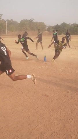 Several people are engaged in a sports activity on a dry, reddish-brown dirt field. They are wearing athletic attire, including soccer jerseys and shorts. Some of the players are wearing the number "50" or "10." There is a blue cone on the ground. The background consists of trees and a light grey sky. The camera is angled, making the scene appear rotated.