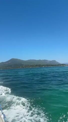 The video shows a view from a boat moving through clear blue water under a bright blue sky. In the distance, a long, green, mountainous coastline is visible. The water is slightly choppy, with white foam trailing behind the boat. The sun is shining, and the overall atmosphere is sunny and serene.