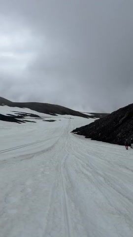 The video shows a group of people hiking on a snowy mountain under a cloudy sky. The terrain is rocky and uneven, with patches of snow and exposed rock. The people are wearing hiking gear and using trekking poles. The video pans across the landscape, showing the vastness of the mountains and the challenging conditions of the hike. The overall impression is one of adventure and exploration in a rugged natural environment.