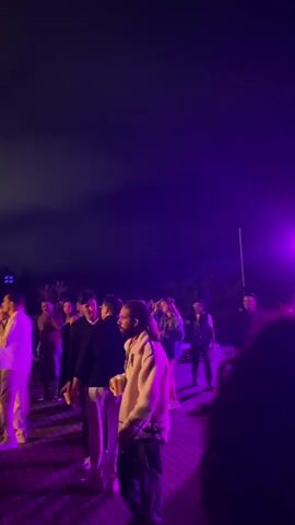 The video shows a nighttime outdoor gathering with a group of people. The scene is illuminated by purple lighting. Several individuals are standing and talking, while some appear to be dancing. The ground is a wooden deck, and trees are visible in the background. The sky is dark, suggesting it is late at night.