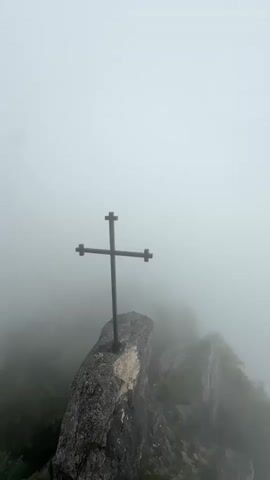 This is a vertical, eye-level shot of a cross on a rocky outcrop, partially obscured by mist. The cross, made of dark metal, has a simple design with four points. It is fixed into a large, grey rock, which itself sits on a precarious ledge. Below and to the right of the main rock, several other rocky formations jut out, also covered in greenery that is softened by the fog. The background is a uniform, hazy white, suggesting dense fog or low clouds that obscure any further landscape.

The weather appears to be foggy or overcast, creating a serene and somewhat isolated atmosphere. The time of day is not explicitly indicated, but the muted light suggests it could be early morning or late afternoon, or simply a heavily clouded day. The scene is set on a mountaintop, given the rocky terrain and the elevated perspective. There are no people visible in the frame.

The prominent object is the cross, a symbol of faith, placed in a high, exposed location. The surrounding rocks and vegetation indicate a natural, possibly mountainous, environment. The overall mood evoked is one of solemnity, solitude, and perhaps spiritual contemplation, enhanced by the misty conditions which create a sense of separation from the world.

No visible text is present in the media file. The location context of Dzirovani, Georgia, places this scene in a specific geographical and potentially cultural setting.
