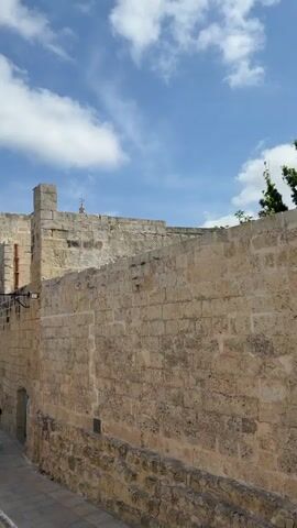 The camera moves along a narrow stone passageway flanked by high stone walls. A church dome is visible over the walls. A white van is parked behind one of the walls. The camera moves towards a lookout point revealing a panoramic view of a sprawling landscape under a blue sky with fluffy white clouds.
