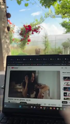 The video begins with a close-up of a laptop screen displaying a YouTube video. The YouTube video features a woman holding a knife, looking intensely at the camera. The laptop is placed on a surface, and behind it, a lush green garden is visible under a clear blue sky. Red flowers, possibly roses, are climbing an archway in the garden. The camera then slowly zooms out, revealing more of the garden with trees, plants, and a greenhouse in the background. The overall scene suggests a peaceful outdoor setting, contrasted with the dramatic imagery on the laptop screen.