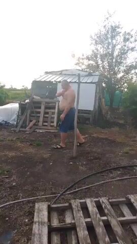 A shirtless man wearing blue shorts and sandals walks through a dirt yard with scattered debris, including wooden pallets, a greenhouse, and a small shed. The man stops and appears to be talking on his phone. He then turns and walks away from the camera towards the shed. The sky is overcast.