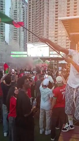 A group of people are gathered outdoors, likely at a sporting event, watching a large screen in the background. The crowd is diverse, with individuals of various ages and ethnicities. Many are wearing red and green, suggesting they are fans of a particular team or country. Some are holding flags, and there is smoke or fog in the air, creating a festive atmosphere. One person in the foreground is standing on a raised platform, waving a green flag and appearing to be leading the cheers. Another person in the foreground is holding a bottle of water and looking towards the screen. The background shows tall buildings, indicating an urban setting. The lighting suggests it is either late afternoon or early evening. The overall mood appears to be energetic and celebratory.