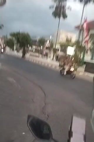 A first-person perspective video shows a person riding a motorcycle on a street. The rider's hand is visible in the lower part of the frame, and a smartphone is mounted on the handlebars, displaying what appears to be a navigation app. Indonesian flags are displayed on poles along the side of the road. Several other motorcycles and a few pedestrians are visible in the distance. The sky is overcast, suggesting it might be late afternoon or early morning. The buildings lining the street appear to be shops or small businesses.