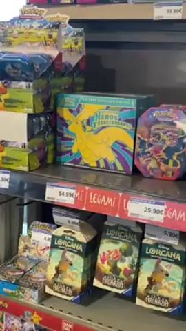 The video shows a store shelf with a variety of collectible trading card games. On the top shelf, there are Pokemon trading cards and sets, including a box with a yellow dragon on the front. Below this, there are stacks of Lorcana cards with Disney characters on the packaging. The middle shelf also has Pokemon cards and One Piece cards. The bottom shelf contains more Lorcana, Pokemon, and Yu-Gi-Oh! cards. All the products have price tags on them. A white tote bag with text in French is visible on the left side of the frame.