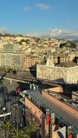 The video shows an aerial view of a city with densely packed buildings in the background. In the foreground, there is a waterfront area with palm trees lining a pedestrian walkway. People are walking along the walkway. A street with cars on it runs parallel to the walkway and water. Buildings of varying heights are interspersed along the waterfront. One building has a sign that reads "EATALY."  The sky is blue with scattered white clouds. The video seems to have been taken during the day.
