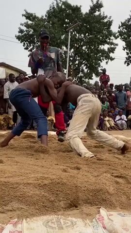In an outdoor setting, the video captures a wrestling match between two young boys on a sand-covered ground. A referee, dressed in a patterned shirt and red shorts, stands nearby observing the match closely. Surrounding the wrestling area is a crowd of spectators of diverse ages and attire. The wrestling ground is marked by what appears to be white sacks positioned around the perimeter. In the background, there are buildings, trees, and a tent-like structure. The sky overhead is overcast, casting a soft light on the scene.