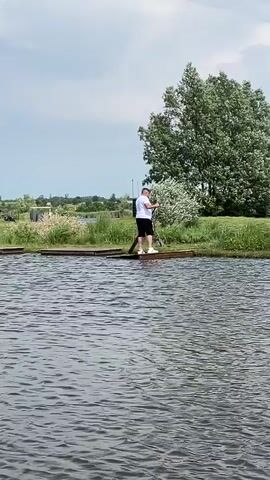 This media file shows a person standing on a wooden dock next to a body of water. The person is wearing a white t-shirt and black shorts, and appears to be holding a long pole, possibly for fishing or stand-up paddleboarding. They are positioned in the middle of the frame, facing away from the camera and towards a large tree with lush green foliage.

The setting is outdoors, with a cloudy sky overhead, suggesting it might be a mild or overcast day. The water in the foreground is rippling, indicating a gentle breeze. In the background, there is more greenery, including tall grass and bushes, with some distant structures or trees visible on the horizon. The overall scene conveys a sense of nature and outdoor recreation. The time of day appears to be daytime, given the lighting. There is no visible text in the media file. The activity seems to be an individual engaging in a water-based activity on a calm day.