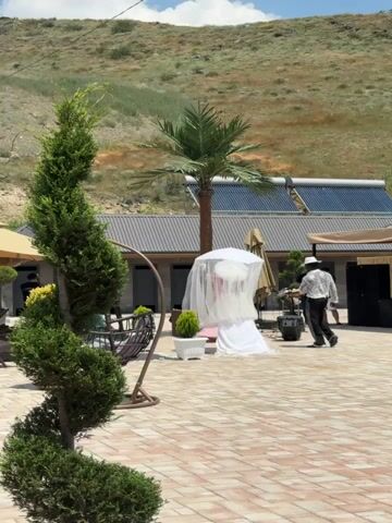 A wedding ceremony appears to be set up in an outdoor courtyard. In the center of the frame, a white wedding dress is draped over a display with a white canopy and a white umbrella overhead, all covered by a sheer white veil. A man in a white shirt with black pants and a white hat, possibly a musician, is standing to the right of the wedding dress, facing it. There is a palm tree behind the wedding dress display. To the left of the wedding dress, a swinging chair with cushions is visible, along with a decorative green shrub. In the background, a building with a grey, corrugated roof and solar panels is situated against a backdrop of a dry, brown hillside. The ground is paved with light-colored square tiles. Two other men are visible in the distance on the right side of the frame, near the building. The lighting suggests it is daytime with clear skies.