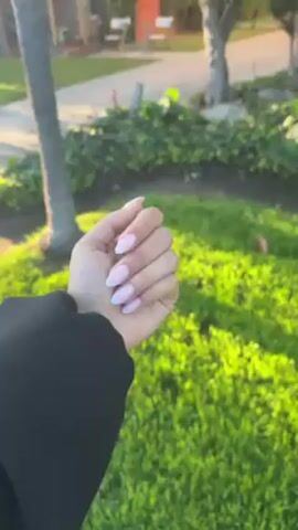 A person wearing a black jacket is holding up their left hand in front of a background of grass, trees, and a building. The person’s hand has manicured nails, painted with a light pink color and light blue tips, and a three-band silver ring is on the ring finger. The hand is posed in two positions, first with fingers together and then with fingers slightly spread. The lighting suggests it is daytime with a bright sun.