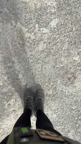 The video begins with a close-up shot of a person's feet wearing dark pants and grey boots, standing on a rough, grey surface. The camera then pans up to reveal the feet of another person, also wearing dark pants and grey boots, standing on a similar surface. The perspective shifts to a wider shot, showing a person from the waist down, wearing dark pants, grey boots, and a tactical vest. The person is standing on a gravelly, concrete ground, with a large concrete structure in the background. A giant mural of a person's face, rendered in shades of grey, is painted on the concrete structure. The structure appears to be part of an industrial or abandoned building, with exposed metal beams and pipes. The camera continues to pan upwards, revealing the full scale of the concrete structure, which is partially enclosed and seems to be a cooling tower or a similar industrial facility. The mural remains the central focus, with the person's feet visible at the bottom of the frame. The environment is desolate and appears to be abandoned, with debris scattered on the ground.