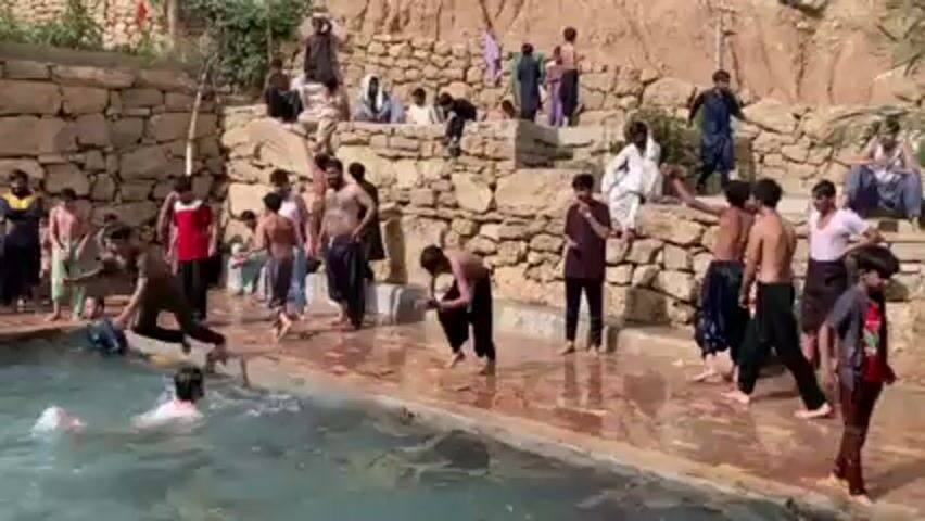 The video shows a group of people swimming and playing in a stone pool. The pool is surrounded by stone walls and terraces, and the environment appears to be sunny and outdoors. Many of the people are young and shirtless, jumping and splashing in the water, while others stand or sit around the pool watching. The video captures the carefree atmosphere and playful interactions between the swimmers.