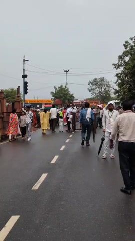 The video shows a street scene with several people walking. The road is wet, indicating it might have rained recently. On the left side of the road, there are some people standing near a red structure with a gate. Further down the road, there is a yellow sign with some text on it, possibly indicating a location. In the center of the road, there is a barrier with people passing through it. On the right side of the road, there are more people, some of whom are holding umbrellas. The sky is overcast, and there is a traffic light on a pole on the left side of the road.