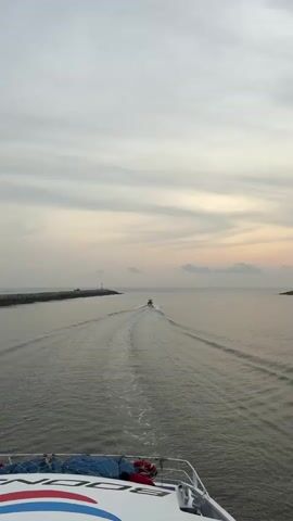 The view from the stern of a boat underway shows a wake behind it, with a smaller boat ahead of it. The ocean is calm under an overcast sky with hints of pink on the horizon. The sun is visible through the clouds. There are short, rocky land masses on either side of the boat, with white lighthouses on each one. The boat has blue, white, and red stripes on its deck, and some equipment secured under netting. A voice can be heard providing information about the boat.