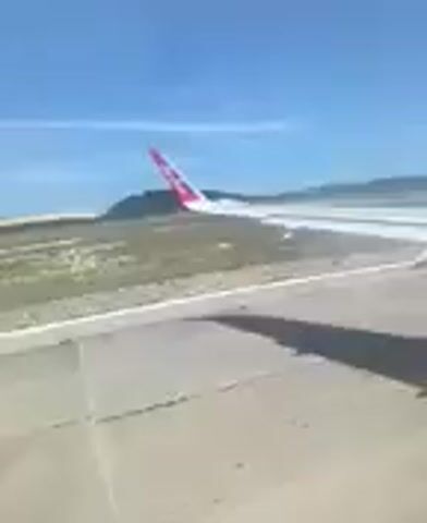 A plane is seen taxiing on a runway. The tail of the plane has a pink logo on it. The sky is blue and clear. There are some hills in the distance. The camera appears to be inside the plane, looking out the window. The plane is moving slowly.
