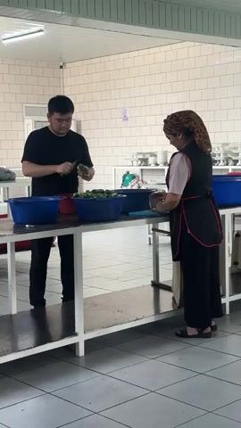 In an indoor setting with tiled walls and floor, two individuals are engaged in food preparation. A man in a black t-shirt and dark pants stands on the left, holding a green vegetable, possibly a cucumber, and a knife. He appears to be peeling or trimming it. In front of him is a large blue tub filled with similar green vegetables. To the right, a woman wearing a headscarf, a light-colored shirt, and a dark apron over dark pants is working at a table. She also holds a green vegetable and a knife, and is in the process of cutting it. In front of her is another blue tub, also containing green vegetables. A third blue tub is visible further to the right. The overall environment suggests a kitchen or food processing area, with shelves and various containers in the background. The lighting appears to be artificial.