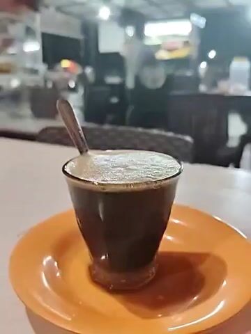 The video shows a glass of coffee on a small orange plate sitting on a white table. The glass has a spoon in it and is full of coffee, with a light-colored foamy top.  The camera pans up to reveal an outdoor setting with tables and chairs in the background. A person wearing a white shirt is seen standing near a food cart in the distance. The scene is set at night under artificial lighting. An Islamic call to prayer can be heard in the background.
