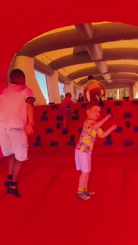The video displays five children and one adult inside a large inflatable obstacle course. The structure features a bright red bouncy floor and climbing walls with blue and black climbing holds. The ceiling is composed of alternating yellow and white inflatable sections, supported by prominent grey inflatable tubes. Three boys are actively climbing the red wall in the background, using the attached holds. In the foreground, a girl wearing a pink t-shirt, grey shorts, and a pink cap stands on the left. A boy in a white t-shirt and white shorts stands beside her. On the right, another boy in a yellow patterned t-shirt and white shorts first stands, then crouches on the red floor. A female adult is partially visible in the far background. The far end of the inflatable structure is open, revealing trees and a clear sky, indicating an outdoor or semi-outdoor location.