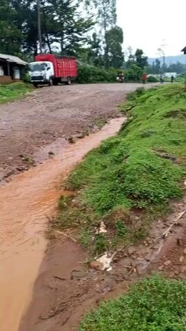 The video shows a muddy road flooded with reddish-brown water. A man in a dark long-sleeved shirt, dark pants, and black boots walks on the left side of the flooded road.  Further up the road, a person wearing a blue top is riding a motorcycle with a large white sack on the back. A red and white truck is parked on the right side of the road. The camera then pans further up the flooded road, focusing on the flowing water.  A white truck approaches from the distance, heading towards the camera. Buildings are visible on both sides of the road. Green grass grows along the edges of the flooded road.