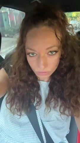 A woman is sitting in a car wearing a white and blue checkered top with a seatbelt on. She has long curly brown hair, partially tied up, and appears to be looking directly at the camera. She adjusts her hair with her right hand. The car is moving, and trees and buildings are visible through the window. Music is playing in the background.
