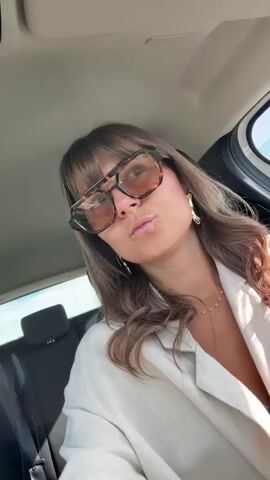 A woman in a car makes faces at the camera. She has brown hair with bangs, and wears brown sunglasses, a beige linen jacket, and large gold earrings. The background shows a car interior and blurry scenery moving past the window. She initially makes a kissing face, then sticks her tongue out playfully.