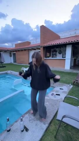 A young person with light brown hair, wearing a black hoodie and grey pants, dances outdoors next to a swimming pool. The person is holding a black object in their right hand and is moving their arms and body rhythmically. The setting appears to be a backyard with a pool, a patio area, and a building with a tiled roof in the background. The sky is cloudy and appears to be dusk. There are several bottles and a pair of sunglasses on the ground near the person.