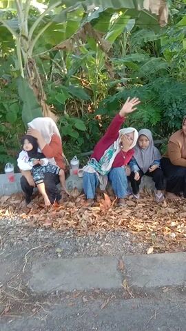 A group of young people, likely students, are gathered outdoors. There are at least four individuals visible in the foreground, all appearing to be female and wearing hijabs. They are seated on the ground, some on fallen leaves and some on concrete. One individual in the front center is waving. In the background, more individuals are visible, some standing and some seated. The environment appears to be a roadside with foliage and trees. The lighting suggests it is daytime.
