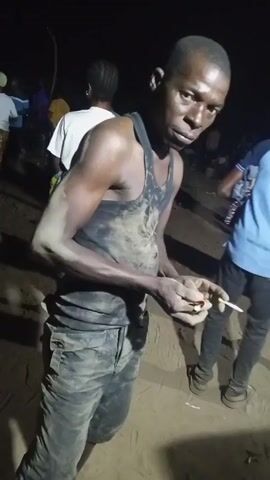 A man is dancing with a cigarette in his hand. He is wearing a black tank top and camouflage pants. He appears to be covered in dirt. Other people can be seen standing around in the background. The lighting suggests it is nighttime.