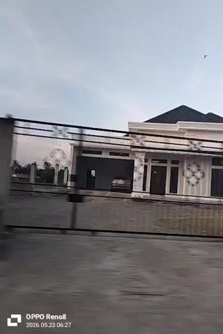 The video shows a view from a moving vehicle, likely a car. Initially, the camera focuses on a modern, single-story house with a black roof and white exterior. As the vehicle progresses, the view shifts to expansive fields, possibly rice paddies, stretching towards a distant tree line and horizon under a partly cloudy sky. The movement of the vehicle blurs the foreground. The video also displays the timestamp and device model on the bottom.