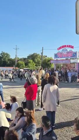The video shows a marching band playing drums in front of a crowd. The drummers are wearing uniforms of white jackets with green trim and berets. They are playing large bass drums, and some of the drummers are raising their drumsticks high in the air before striking the drums. The crowd is watching the band, and some people are taking pictures. The scene is taking place on a street, and there are trees and buildings in the background. The weather appears to be sunny.