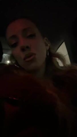 The video shows a woman in a car at night. The interior of the car is dark, with only a little bit of ambient light coming in from outside. The woman is wearing hoop earrings and a dark red top. She is looking directly at the camera and talking or singing along to music. The video appears to be a selfie-style recording.