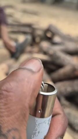 A close-up shot shows a hand holding a white and silver lighter. The lighter is lit, with a small orange flame flickering from its top. In the background, a pile of chopped wood is visible, suggesting the setting is outdoors and likely a place where a fire is intended. The lighting appears to be natural, possibly indicating daytime. The image has a slightly grainy quality. There is some text visible on the lighter, but it is mostly obscured by the hand and the angle.