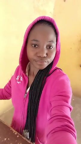 A woman wearing a pink hooded sweatshirt is seen from the chest up, standing indoors with a yellow wall behind her. She has dark skin, braids, and a serious expression. She is holding a phone or camera in her right hand, taking a selfie. The background is a plain interior space.