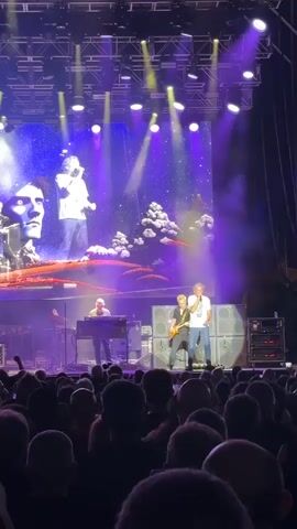 The video captures a live concert performance from the perspective of the audience. A large LED screen dominates the background, displaying abstract imagery of starry skies, fantastical clouds, and stylized portraits of male faces. On stage, a band performs. In the foreground, an audience is visible, their backs to the camera, with some holding up phones to record. The stage is lit with purple and white spotlights, and smoke or haze is present, creating atmospheric effects. A male singer in a white shirt and blue jeans stands center stage, singing into a microphone. To his left, another male musician plays a guitar. Further left, a third musician is seated at a keyboard. Two large speaker cabinets are positioned behind the singer and guitarist. The overall atmosphere appears to be energetic and immersive.