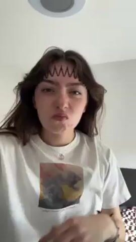 A young woman is seen close-up, centered in the frame, in what appears to be a bedroom. The walls and ceiling are white, and a round light fixture is visible in the ceiling. The woman has long, dark hair with light brown highlights and wears a white T-shirt with a graphic print on the front. She is wearing multiple bracelets on both wrists and a necklace with a small pendant. Initially, she has temporary black markings on her forehead, resembling a jagged design. With her hands, she manipulates her hair to cover the markings, transforming it into a braided style along her hairline. She looks at the camera with a neutral expression throughout the video. The background shows a glimpse of what appears to be bedding.
